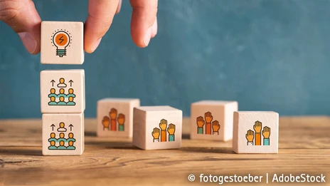 Auf einer Holztischplatte vor einem blauen Hintergrund liegen verschiedene Holzklötzchen mit verschiedenen Aufdrucken wie Händen und Personen; von einer Hand werden drei Klötzchen übereinander gestapelt.